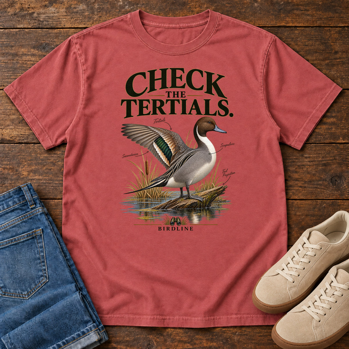 Crimson Check the Tertials Duck T-Shirt with a duck raising one wing beside reeds and water; front flat lay on wood.
