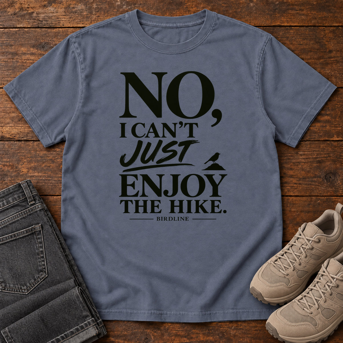 Denim No I Can't Just Enjoy the Hike T-Shirt with bold black lettering and a small bird silhouette, front flat lay on wood.