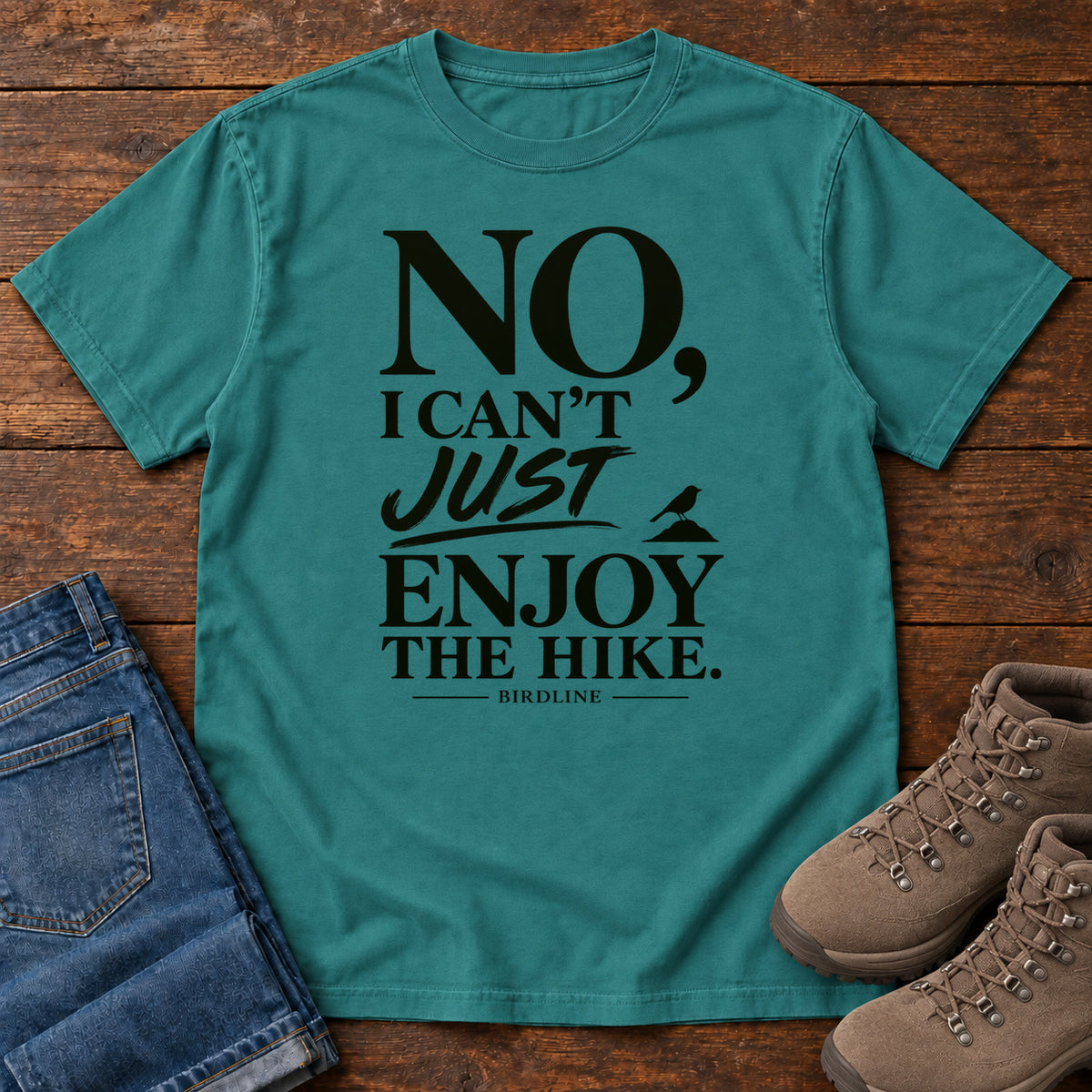 Emerald No I Can't Just Enjoy the Hike T-Shirt with bold black lettering and a small bird silhouette, front flat lay on wood.