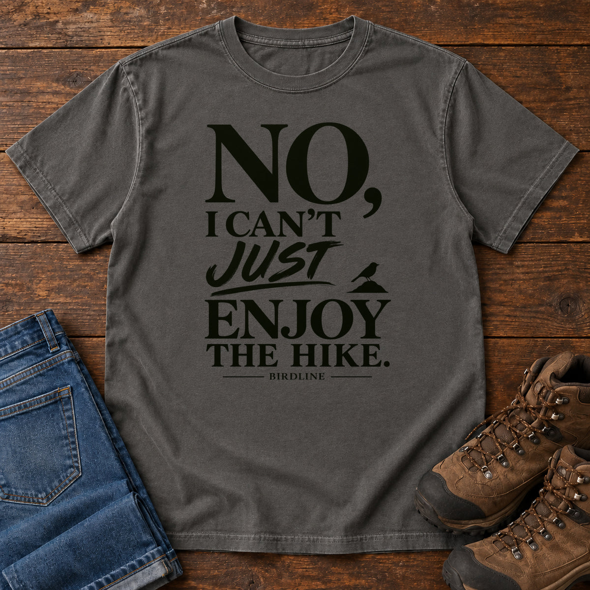 Pepper No I Can't Just Enjoy the Hike T-Shirt with bold black lettering and a small bird silhouette, front flat lay on wood.