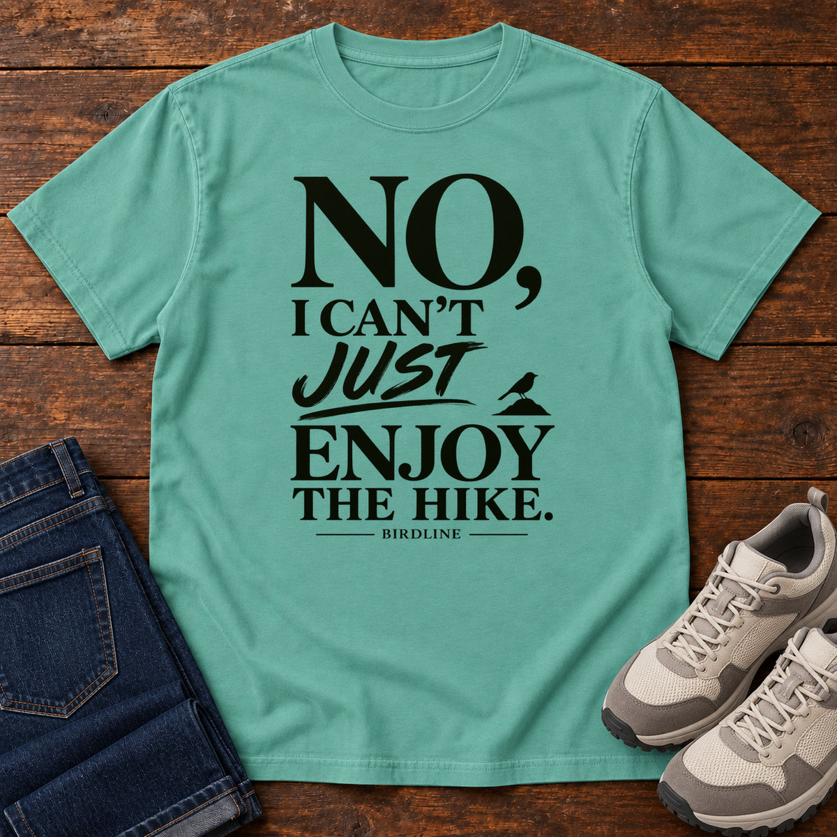 Seafoam No I Can't Just Enjoy the Hike T-Shirt with bold black lettering and a small bird silhouette, front flat lay on wood.