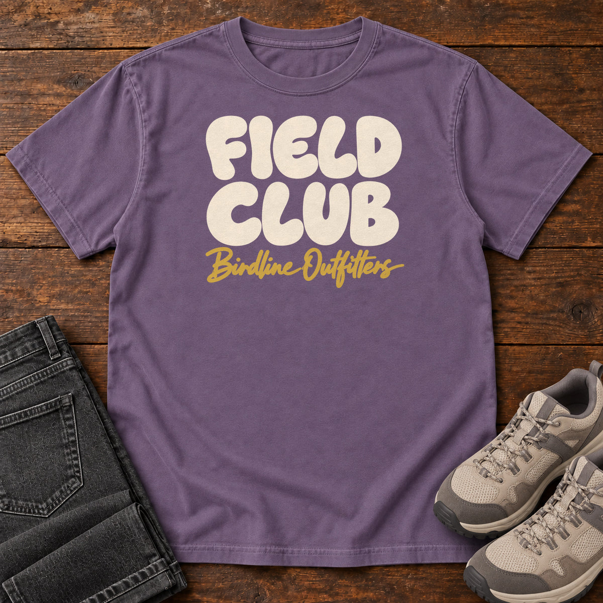 Grape Field Club T-shirt with rounded cream Field Club lettering above gold Birdline Outfitters script, front flat lay on wood.