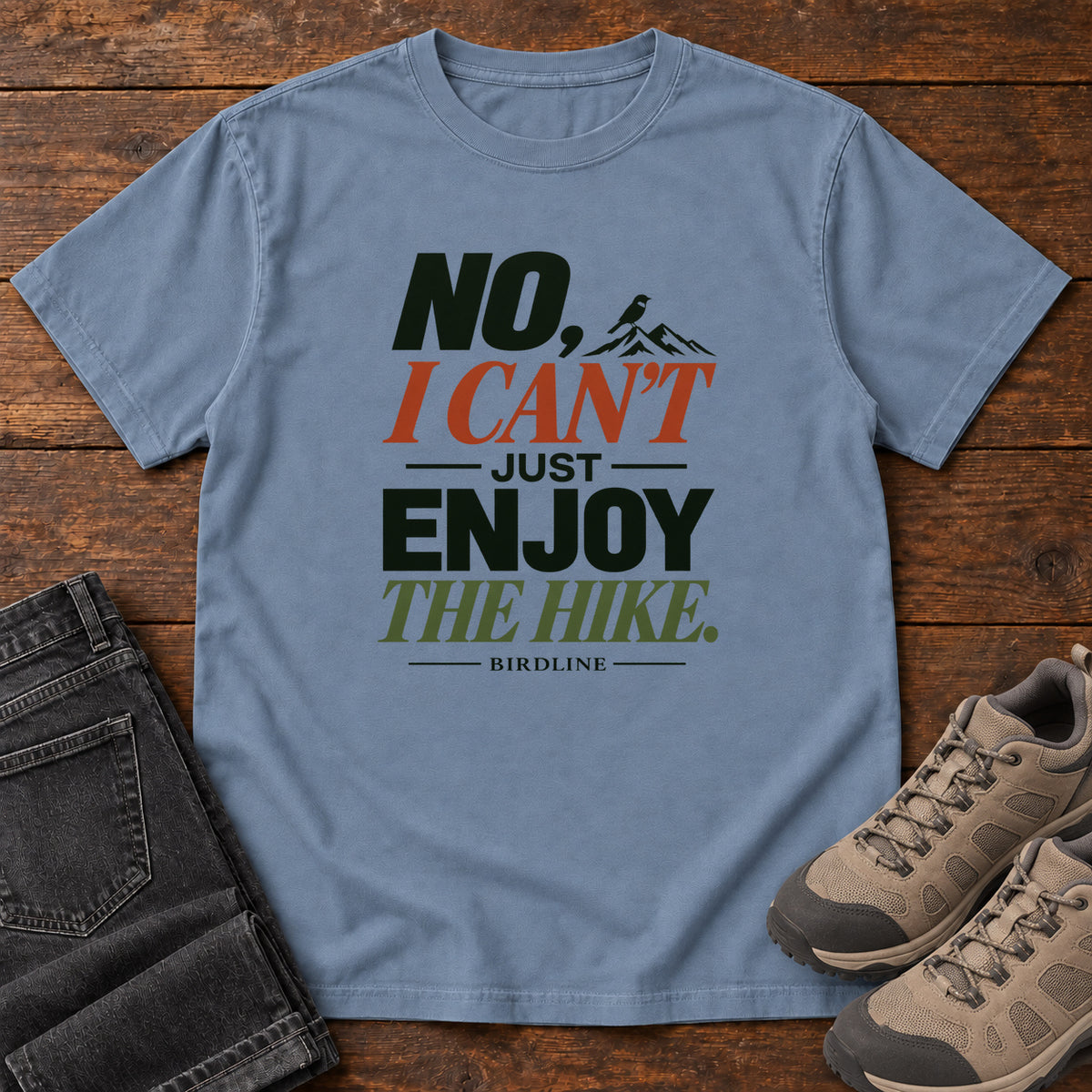 No I Can't Just Enjoy the Hike T-Shirt – Color Block in Blue Jean, front laid flat on wood, featuring stacked green and rust slogan lettering and a small mountain graphic.
