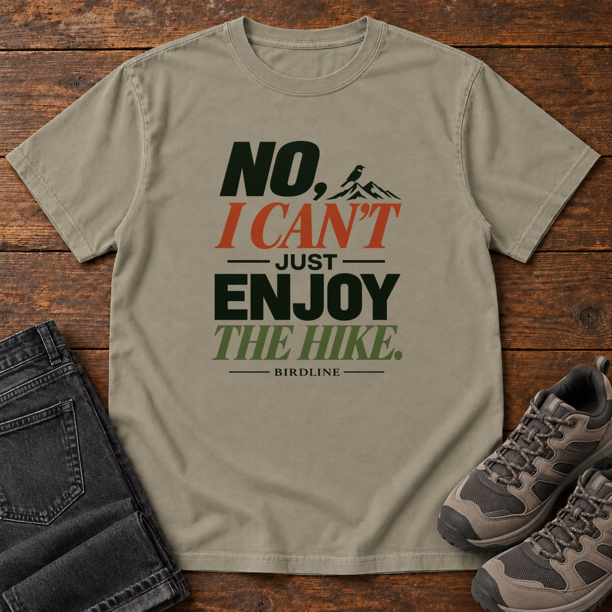 No I Can't Just Enjoy the Hike T-Shirt – Color Block in Sandstone, front laid flat on wood, featuring stacked green and rust slogan lettering and a small mountain graphic.