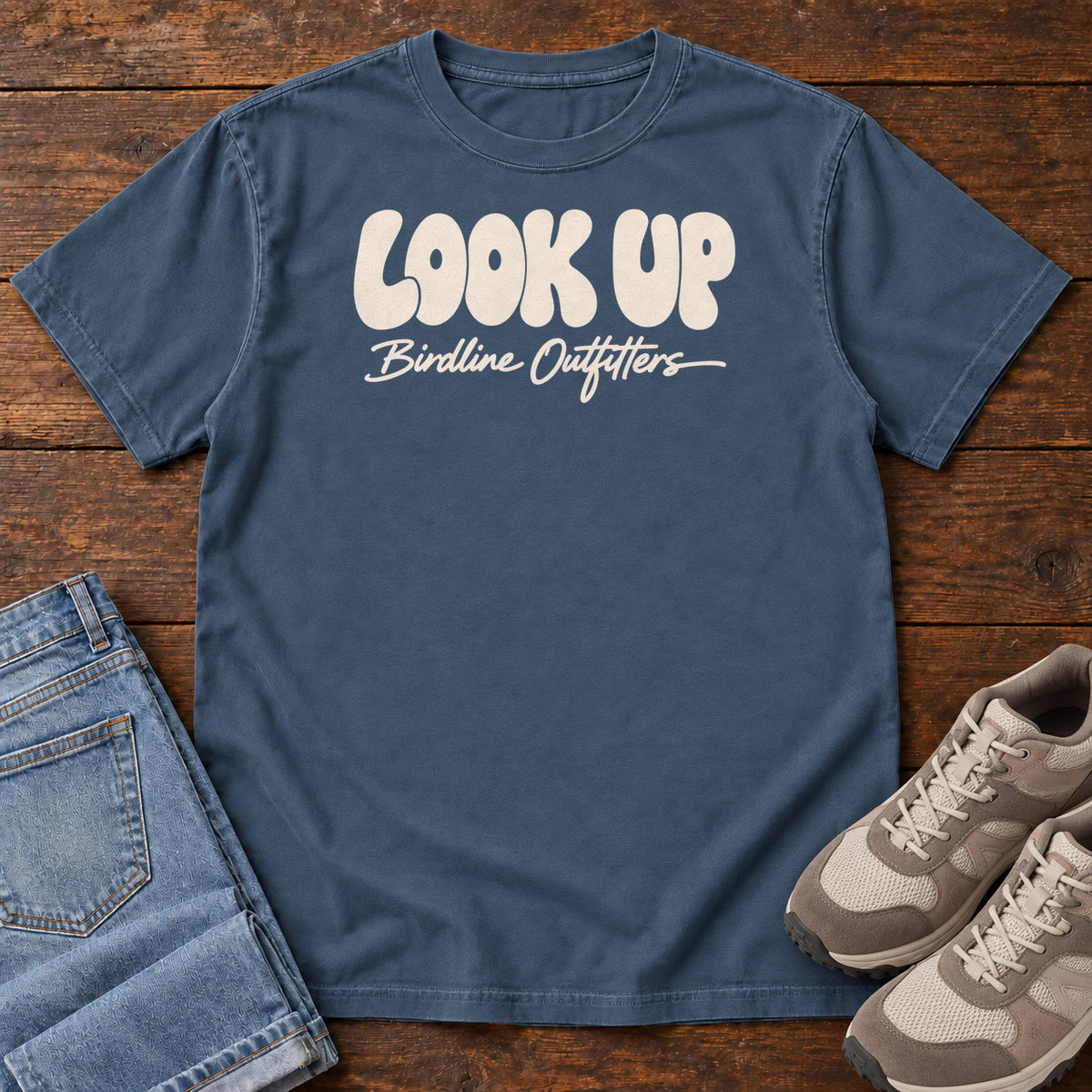 Look Up T-shirt in Midnight, front laid flat on wood, featuring large rounded cream LOOK UP lettering above Birdline Outfitters script.