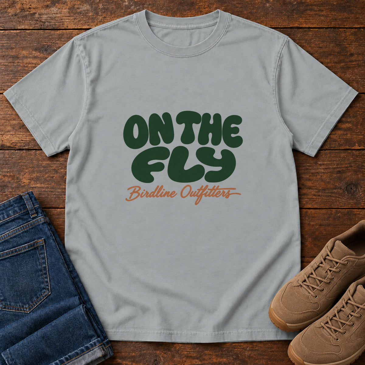 Granite On the Fly T-shirt with rounded dark green On the Fly lettering above rust-orange Birdline Outfitters script, front flat lay on wood.