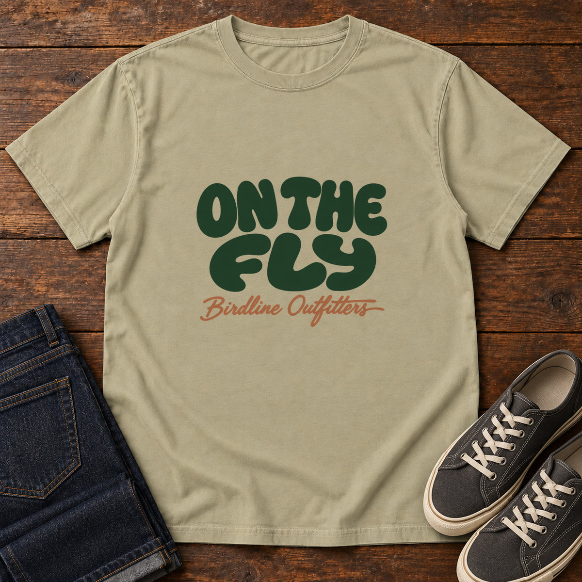 Khaki On the Fly T-shirt with rounded dark green On the Fly lettering above rust-orange Birdline Outfitters script, front flat lay on wood.