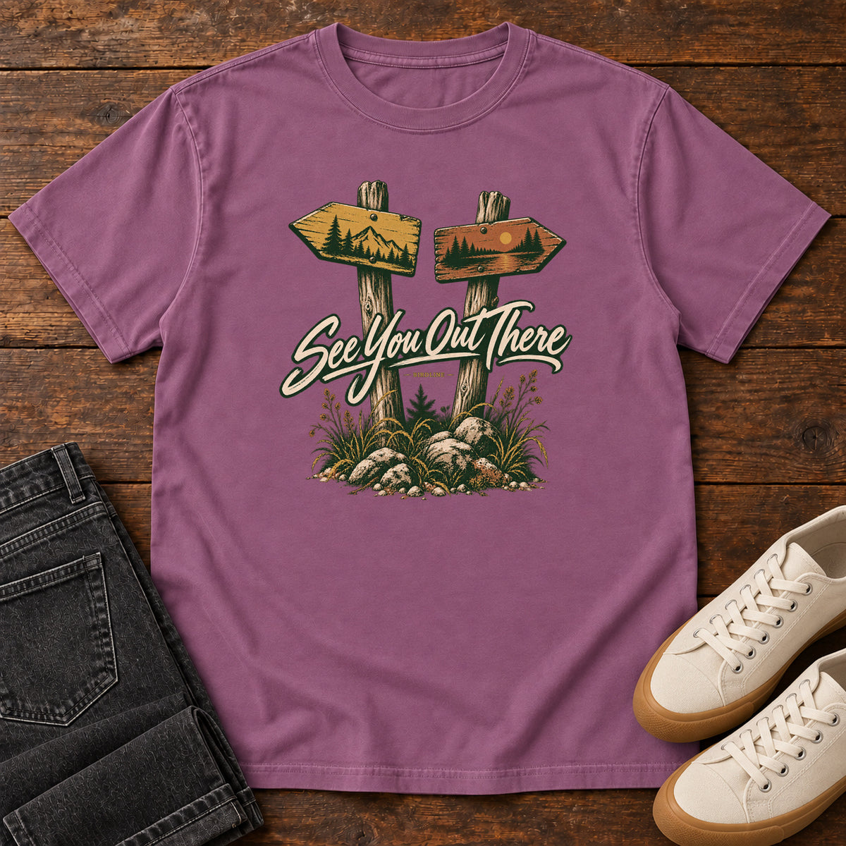 Berry See You Out There T-Shirt with two illustrated trail signposts, rocks, grass and cream script, front flat lay on wood.