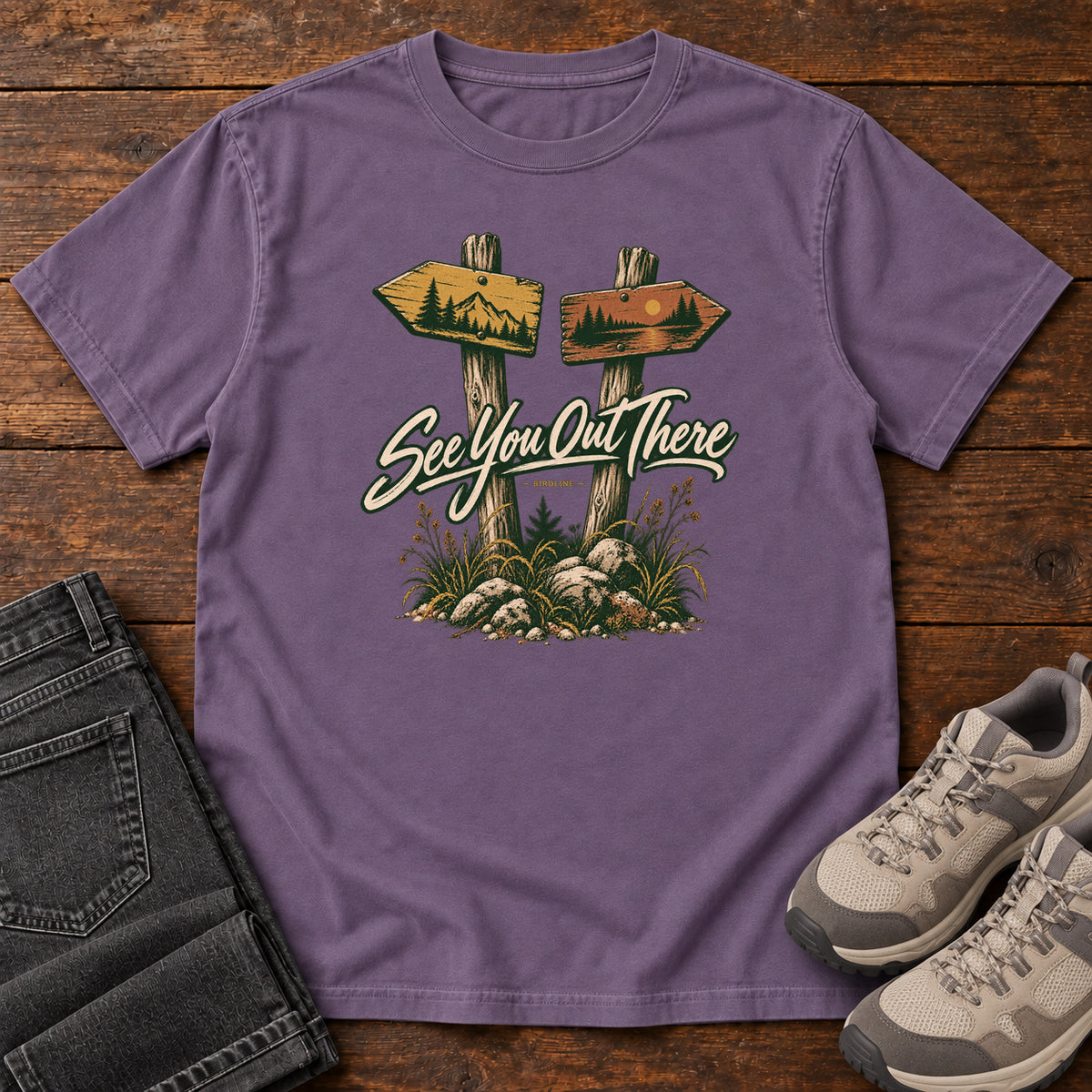 Grape See You Out There T-Shirt with two illustrated trail signposts, rocks, grass and cream script, front flat lay on wood.