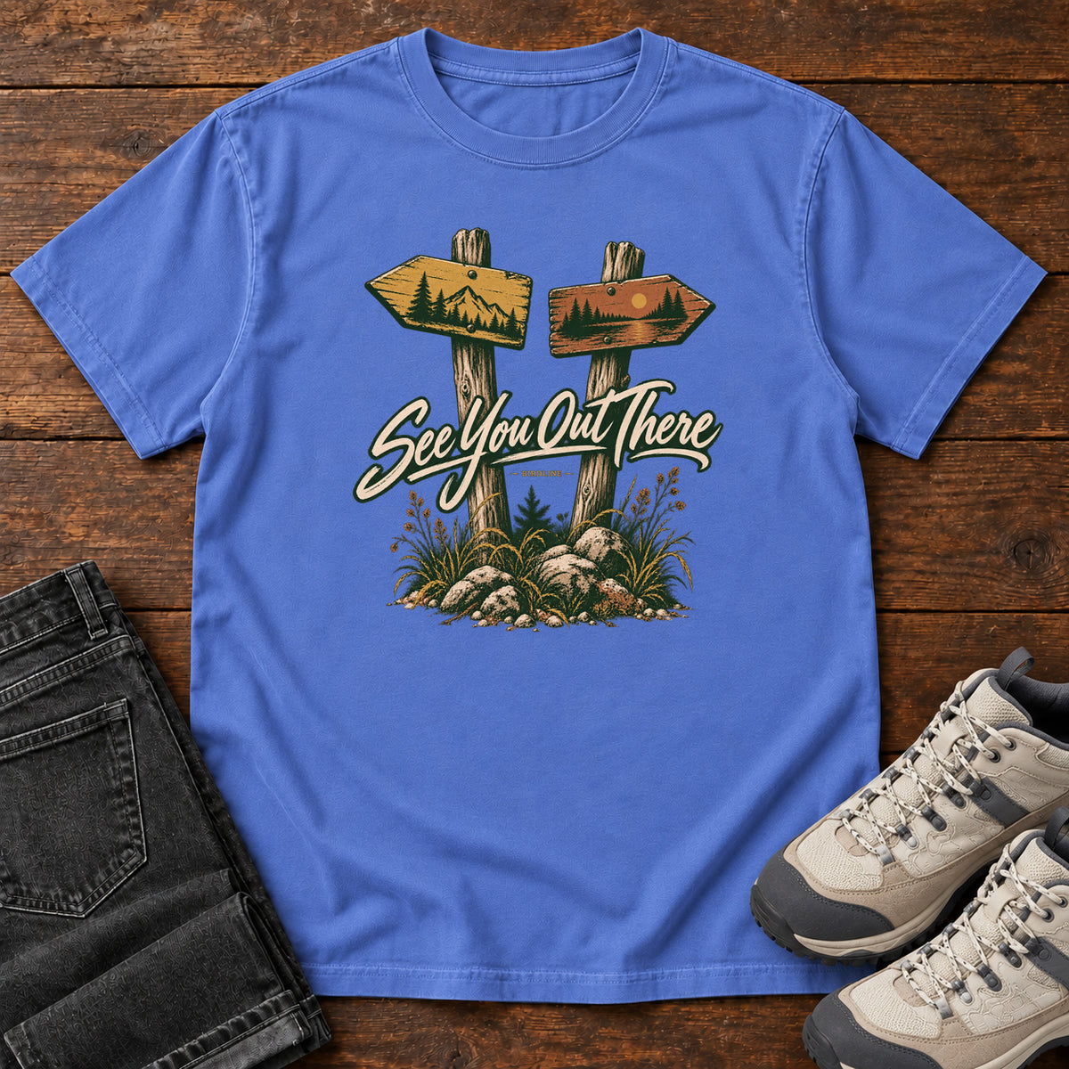 Mystic Blue See You Out There T-Shirt with two illustrated trail signposts, rocks, grass and cream script, front flat lay on wood.