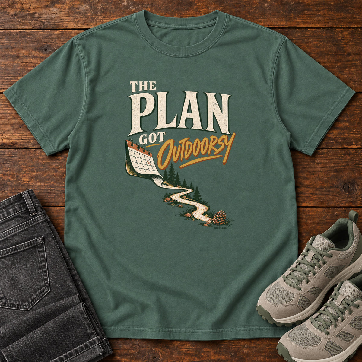 Blue Spruce The Plan Got Outdoorsy T-Shirt with cream-and-gold lettering above a calendar turning into a winding trail, front flat lay on wood.