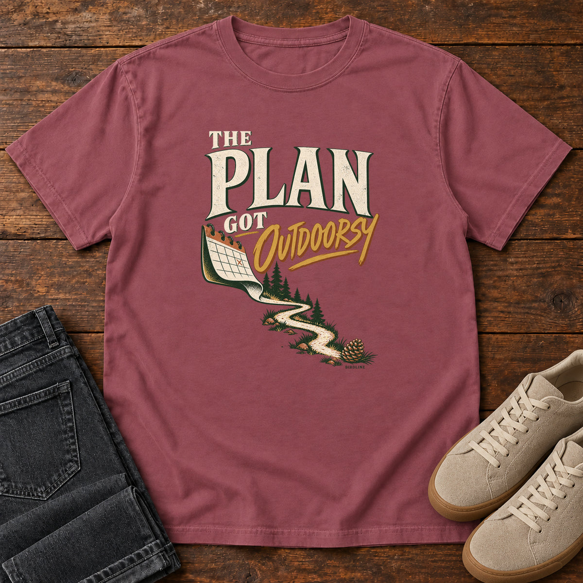 Brick The Plan Got Outdoorsy T-Shirt with cream-and-gold lettering above a calendar turning into a winding trail, front flat lay on wood.