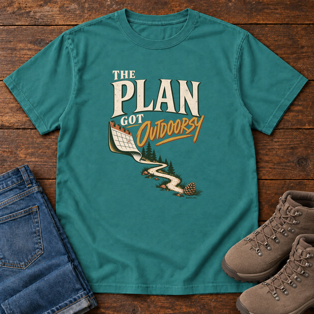 Emerald The Plan Got Outdoorsy T-Shirt with cream-and-gold lettering above a calendar turning into a winding trail, front flat lay on wood.