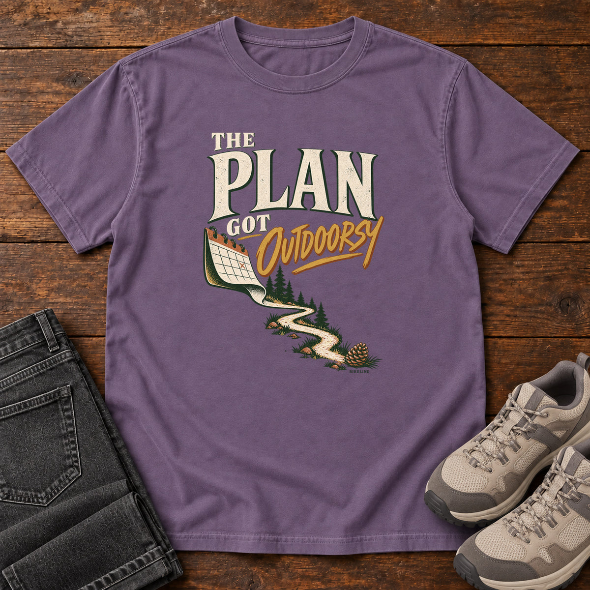 Grape The Plan Got Outdoorsy T-Shirt with cream-and-gold lettering above a calendar turning into a winding trail, front flat lay on wood.