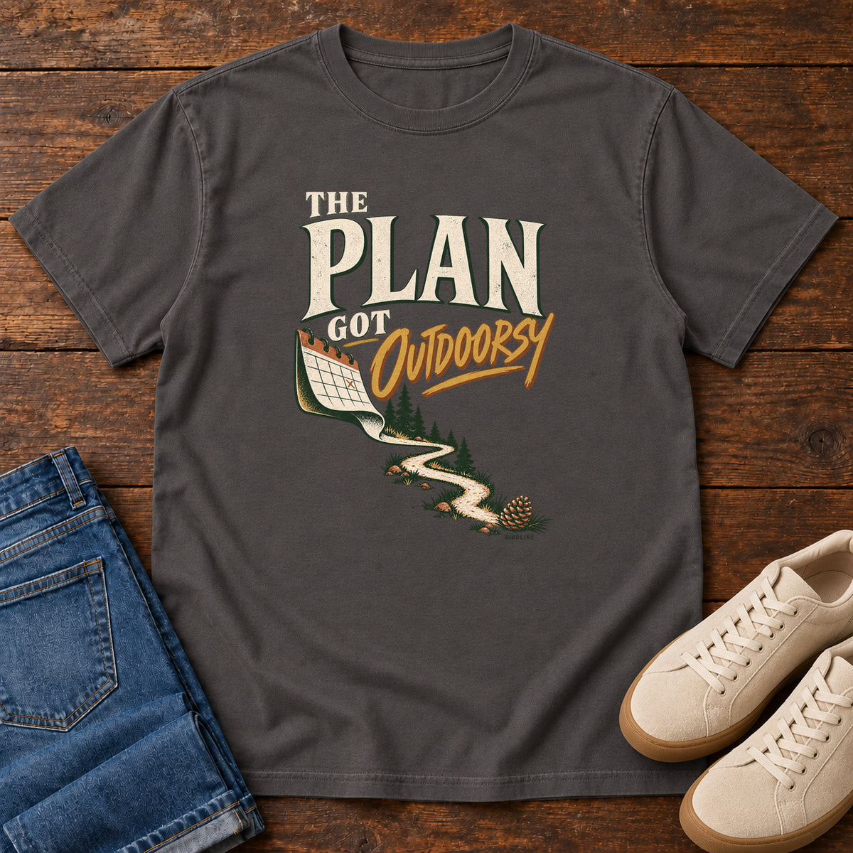 Graphite The Plan Got Outdoorsy T-Shirt with cream-and-gold lettering above a calendar turning into a winding trail, front flat lay on wood.