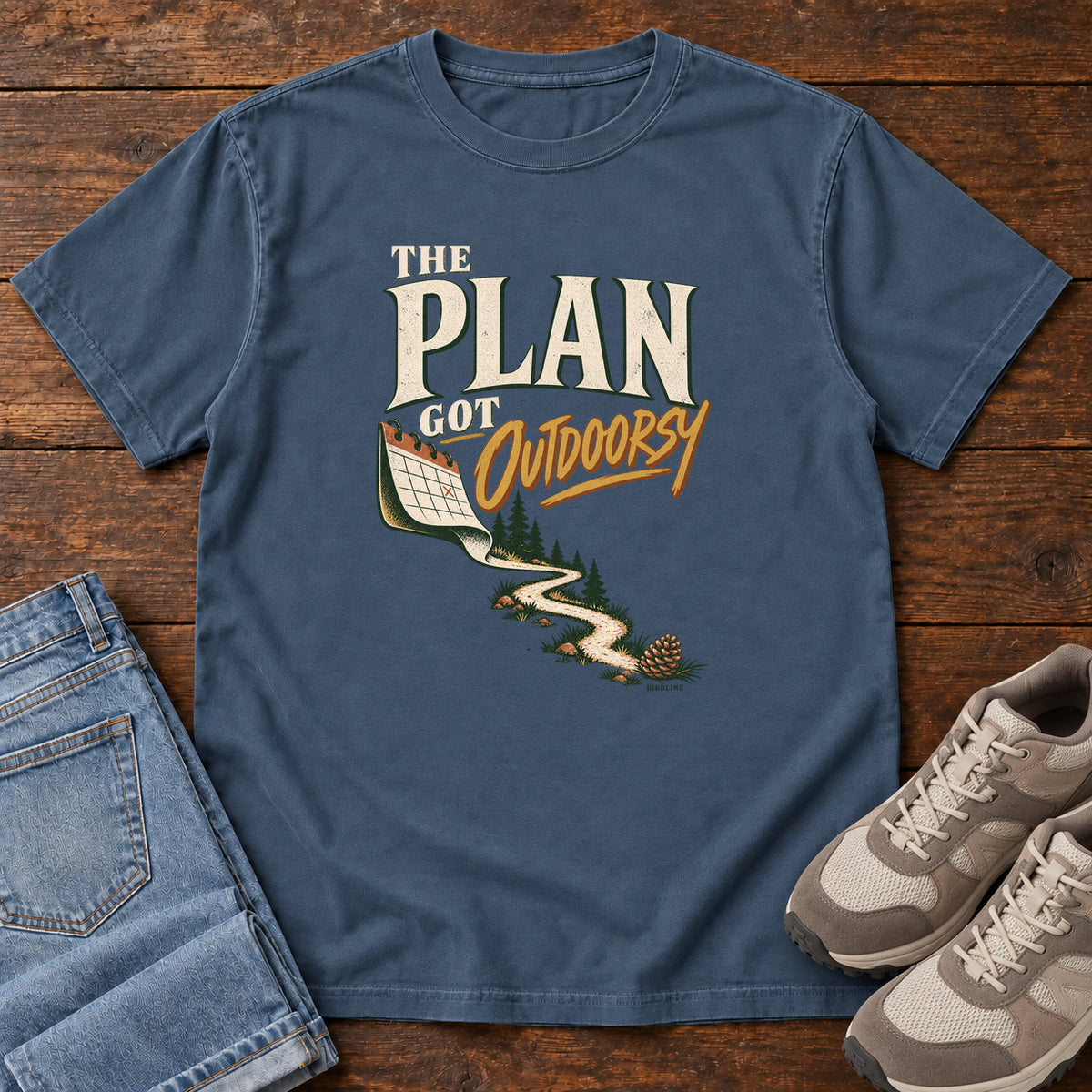 Midnight The Plan Got Outdoorsy T-Shirt with cream-and-gold lettering above a calendar turning into a winding trail, front flat lay on wood.