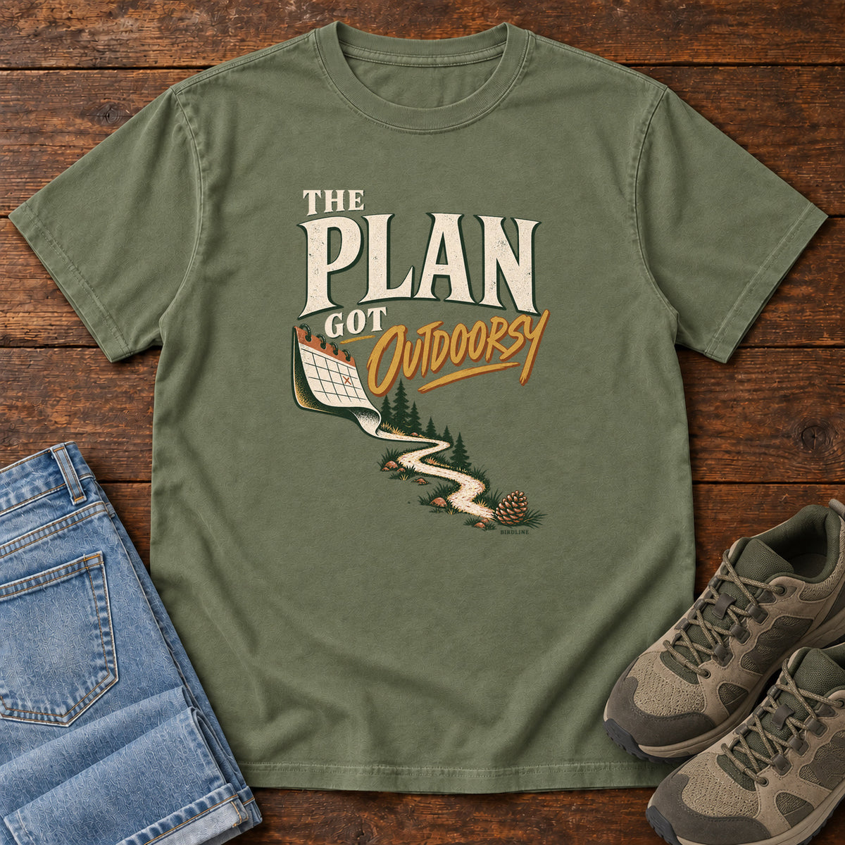 Moss The Plan Got Outdoorsy T-Shirt with cream-and-gold lettering above a calendar turning into a winding trail, front flat lay on wood.