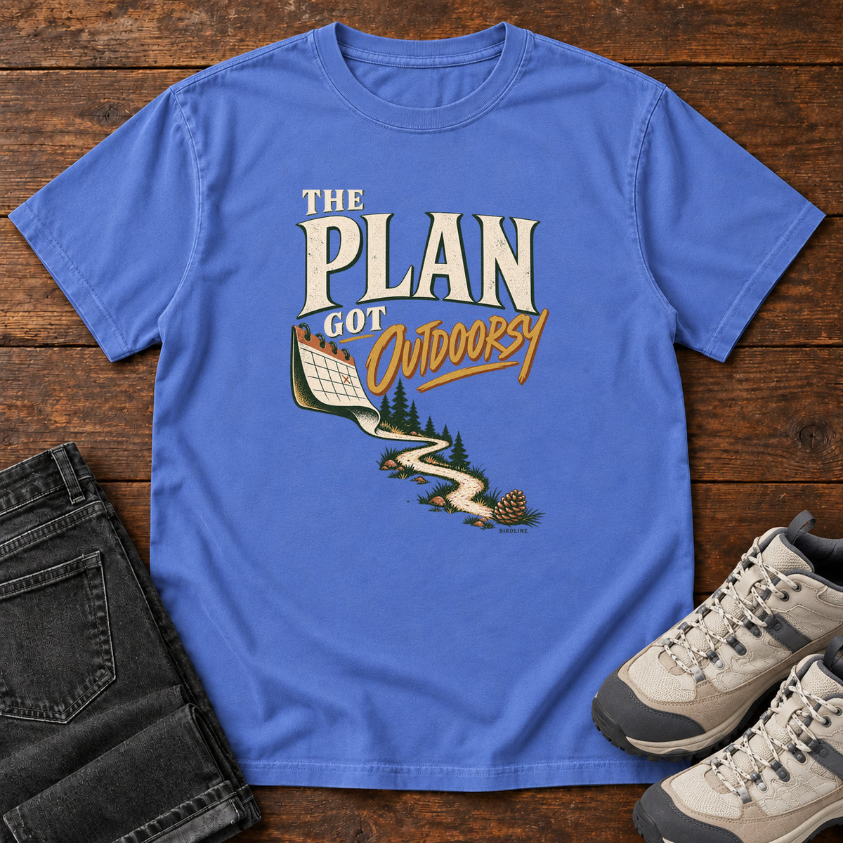 Mystic Blue The Plan Got Outdoorsy T-Shirt with cream-and-gold lettering above a calendar turning into a winding trail, front flat lay on wood.