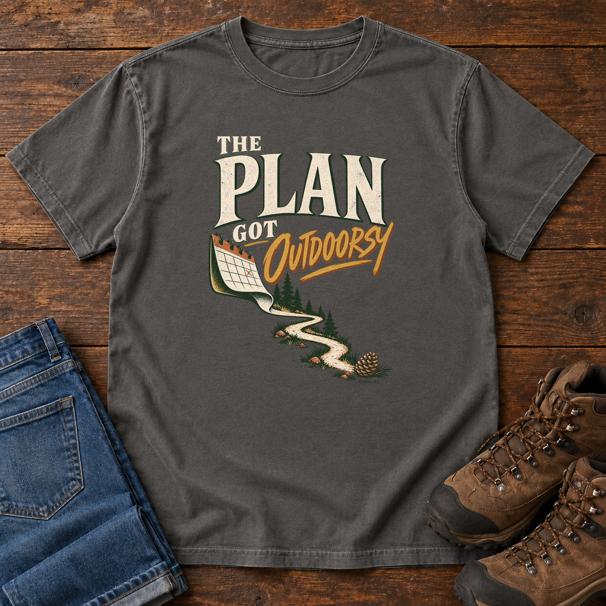 Pepper The Plan Got Outdoorsy T-Shirt with cream-and-gold lettering above a calendar turning into a winding trail, front flat lay on wood.