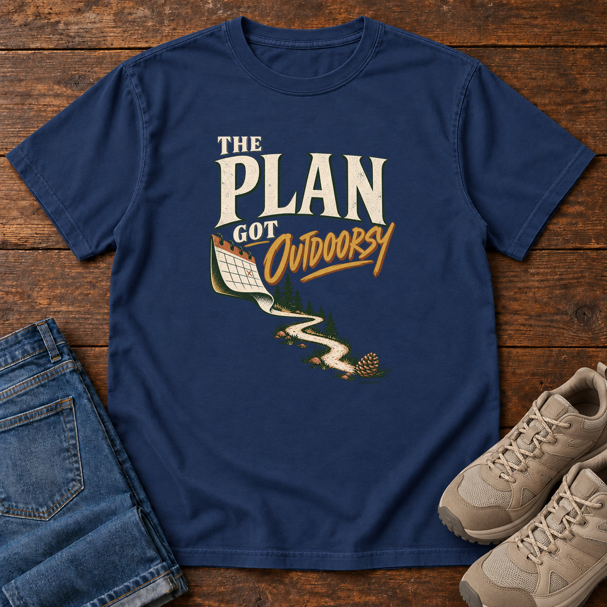 True Navy The Plan Got Outdoorsy T-Shirt with cream-and-gold lettering above a calendar turning into a winding trail, front flat lay on wood.