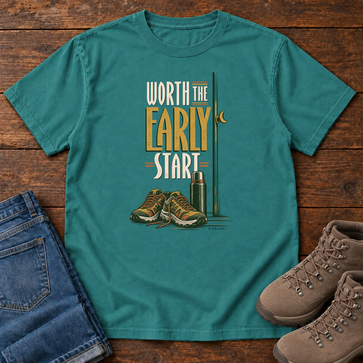 Emerald Worth the Early Start T-Shirt with hiking shoes, a thermos, a walking pole and a crescent moon; front flat lay on wood.
