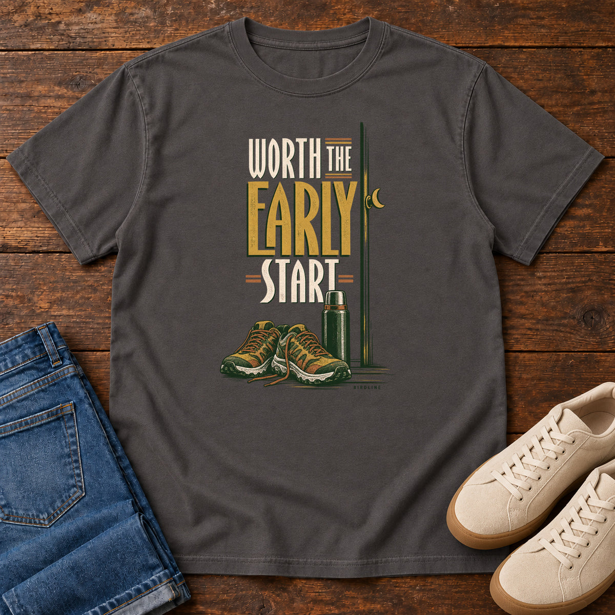 Graphite Worth the Early Start T-Shirt with hiking shoes, a thermos, a walking pole and a crescent moon; front flat lay on wood.