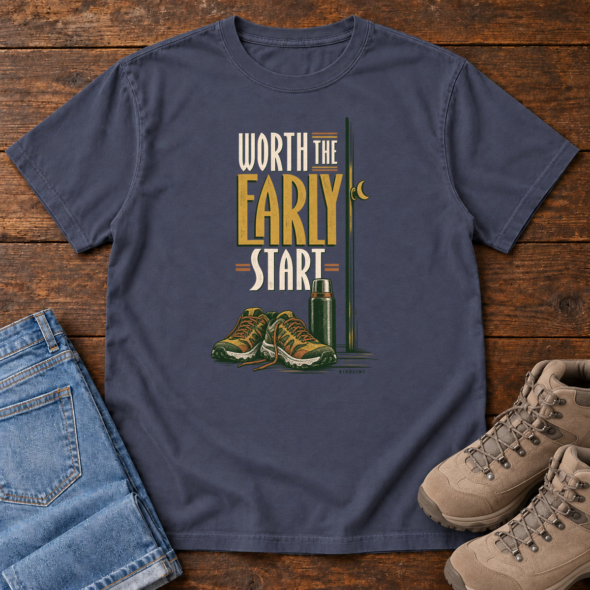 Navy Worth the Early Start T-Shirt with hiking shoes, a thermos, a walking pole and a crescent moon; front flat lay on wood.