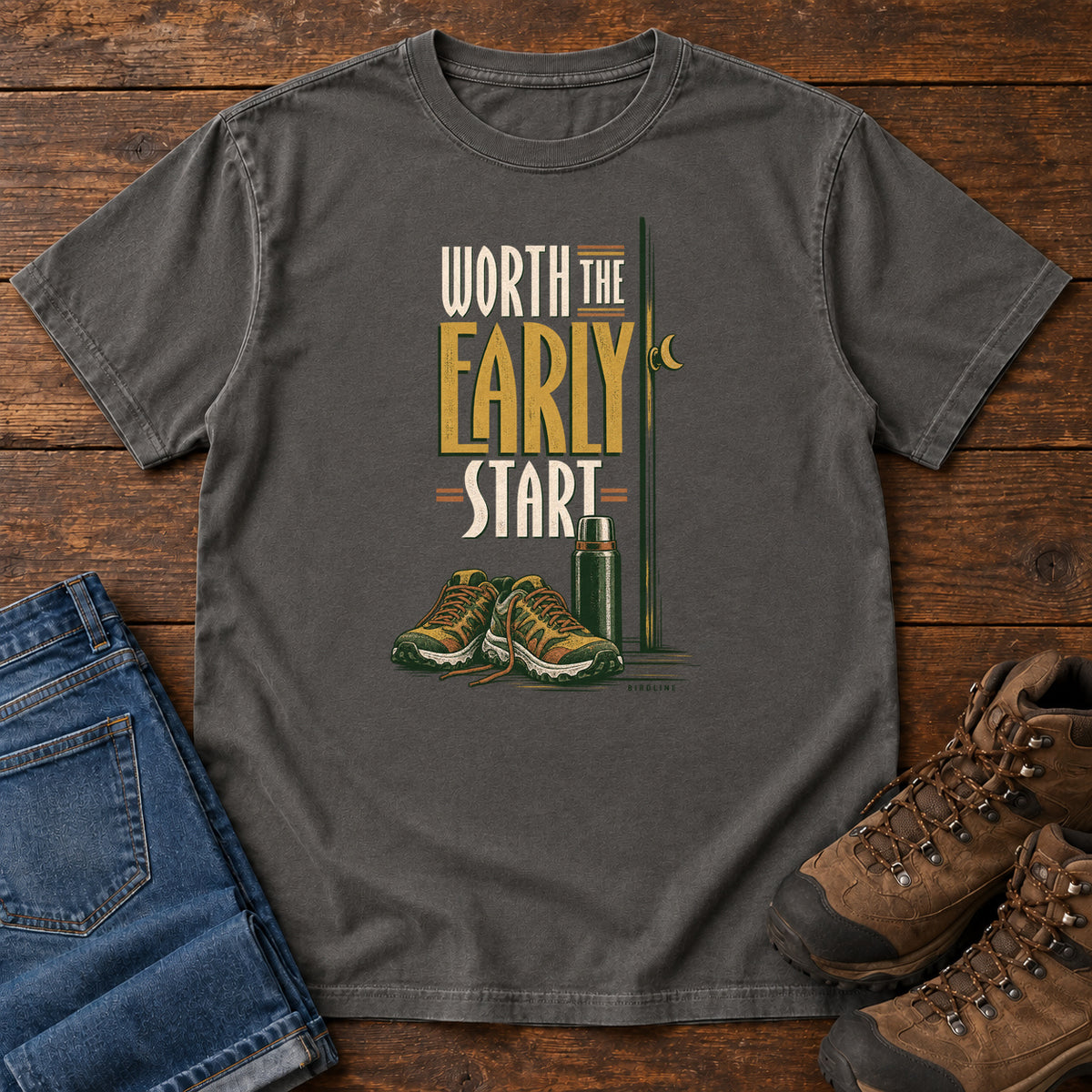 Pepper Worth the Early Start T-Shirt with hiking shoes, a thermos, a walking pole and a crescent moon; front flat lay on wood.
