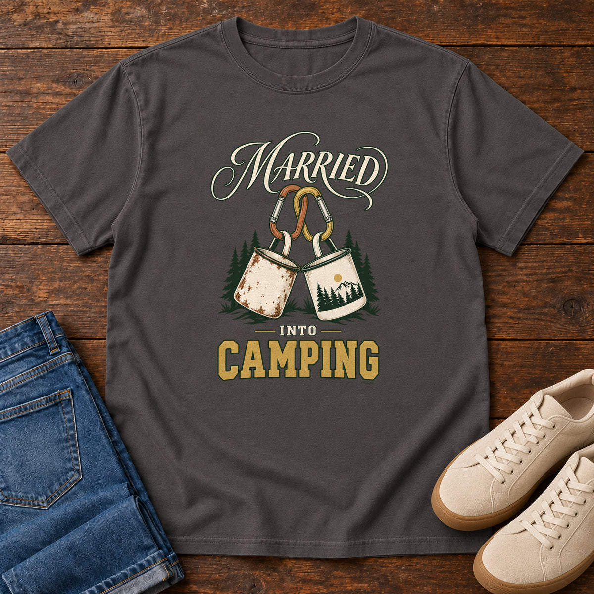 Graphite Married into Camping T-Shirt with two camp mugs hanging from carabiners between cream and gold lettering, front flat lay on wood.