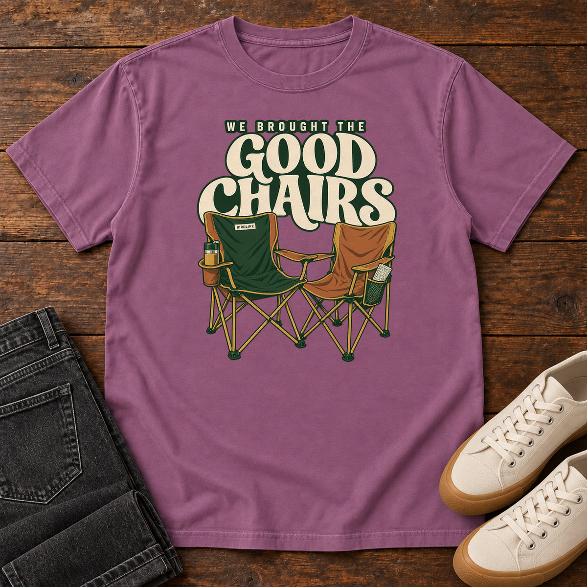 We Brought the Good Chairs T-Shirt in Berry, front laid flat on wood, featuring green and brown folding camp chairs beneath large cream slogan lettering.