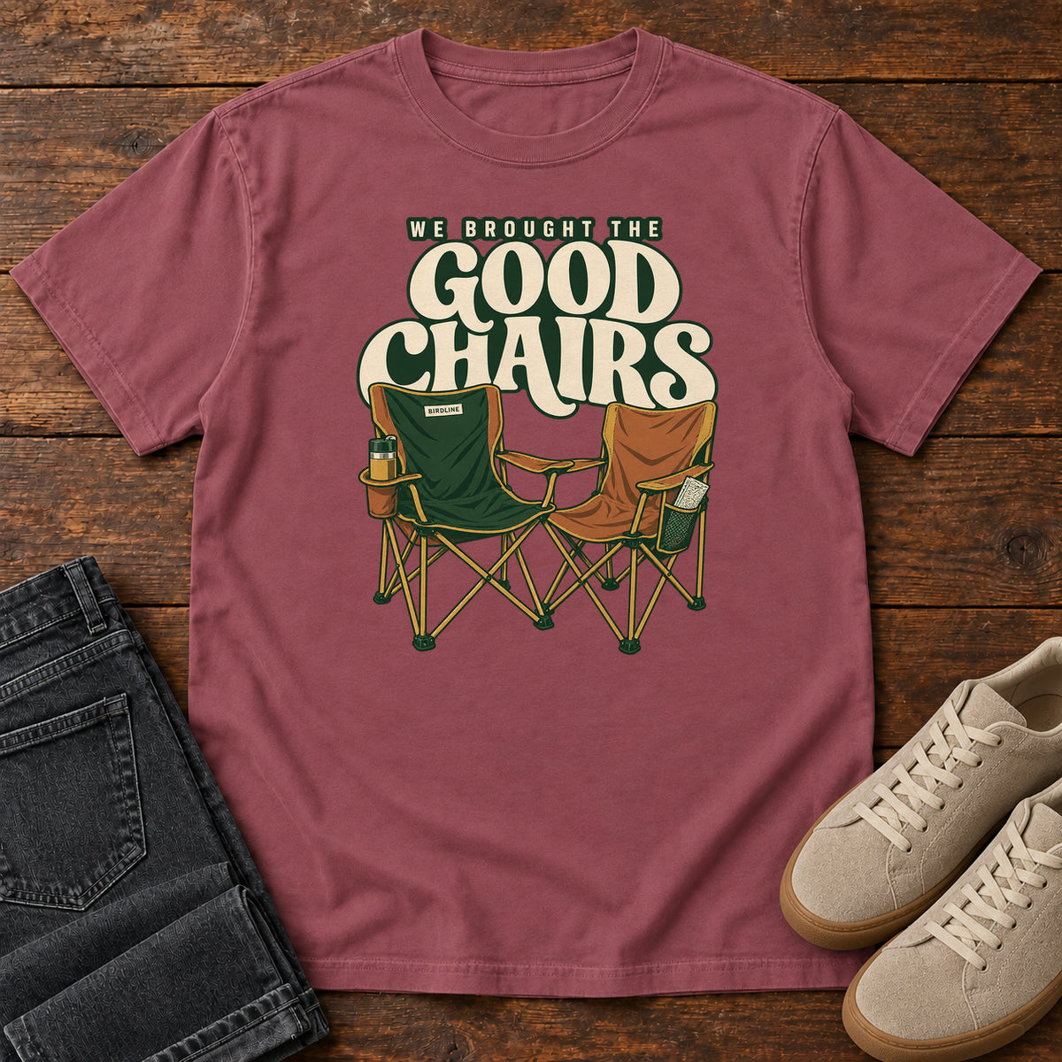 We Brought the Good Chairs T-Shirt in Brick, front laid flat on wood, featuring green and brown folding camp chairs beneath large cream slogan lettering.