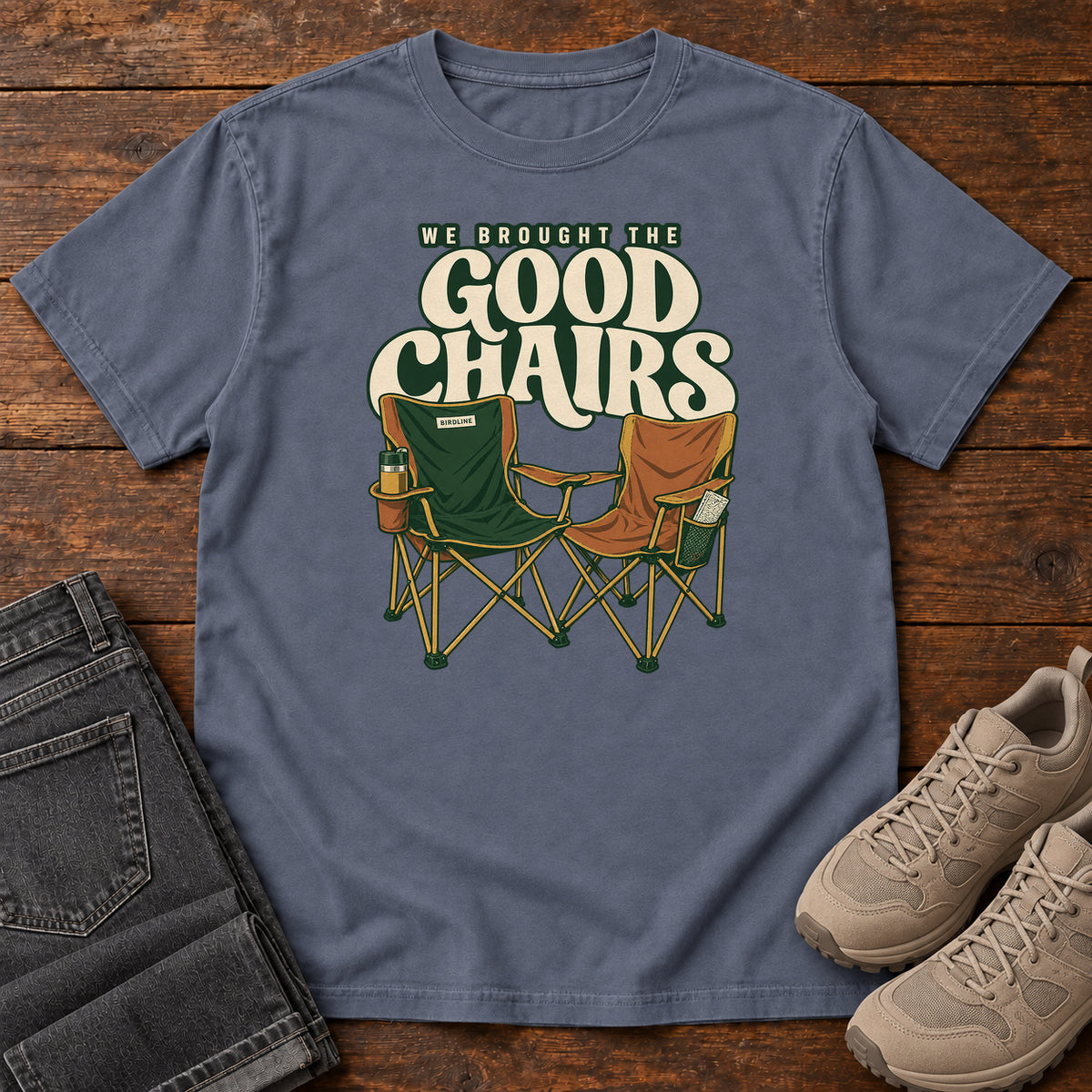 We Brought the Good Chairs T-Shirt in Denim, front laid flat on wood, featuring green and brown folding camp chairs beneath large cream slogan lettering.