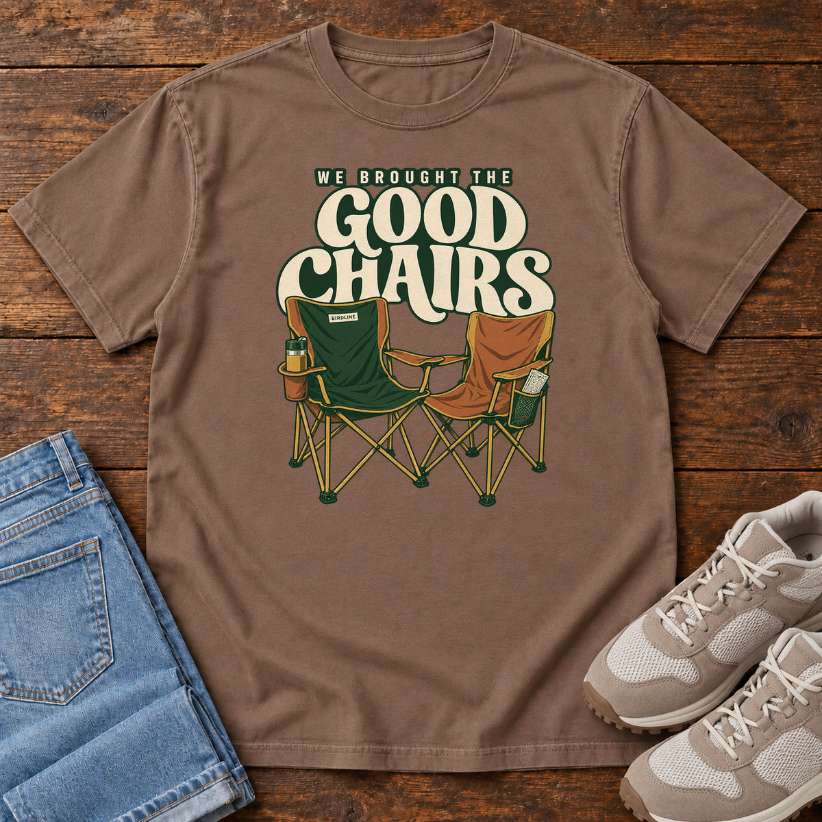 We Brought the Good Chairs T-Shirt in Espresso, front laid flat on wood, featuring green and brown folding camp chairs beneath large cream slogan lettering.