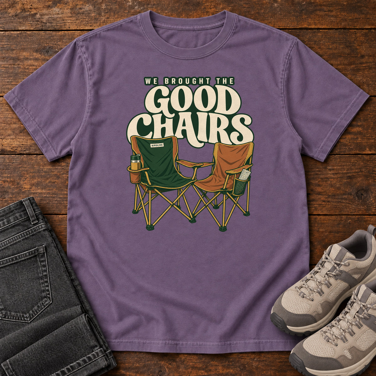 We Brought the Good Chairs T-Shirt in Grape, front laid flat on wood, featuring green and brown folding camp chairs beneath large cream slogan lettering.