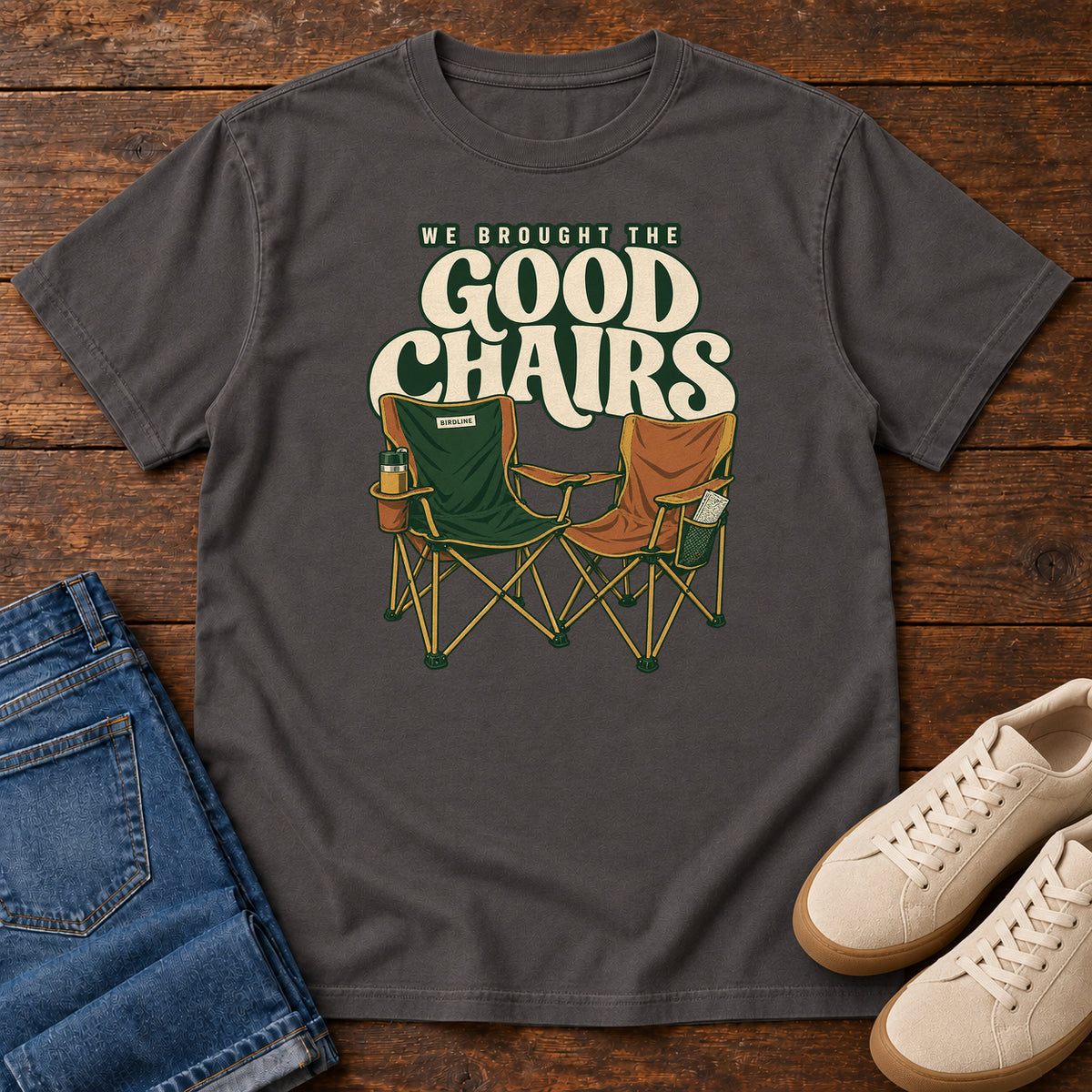 We Brought the Good Chairs T-Shirt in Graphite, front laid flat on wood, featuring green and brown folding camp chairs beneath large cream slogan lettering.