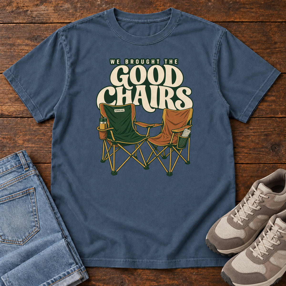 We Brought the Good Chairs T-Shirt in Midnight, front laid flat on wood, featuring green and brown folding camp chairs beneath large cream slogan lettering.
