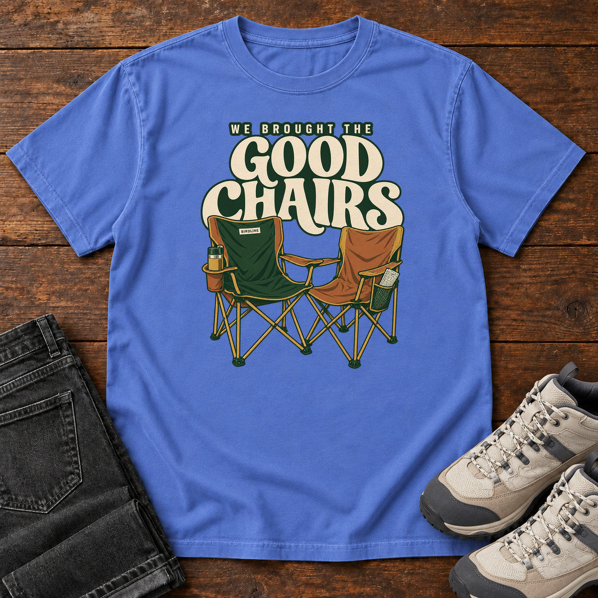 We Brought the Good Chairs T-Shirt in Mystic Blue, front laid flat on wood, featuring green and brown folding camp chairs beneath large cream slogan lettering.
