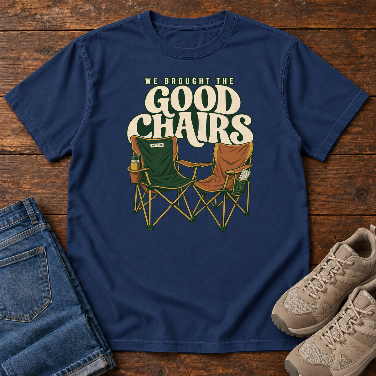We Brought the Good Chairs T-Shirt in True Navy, front laid flat on wood, featuring green and brown folding camp chairs beneath large cream slogan lettering.