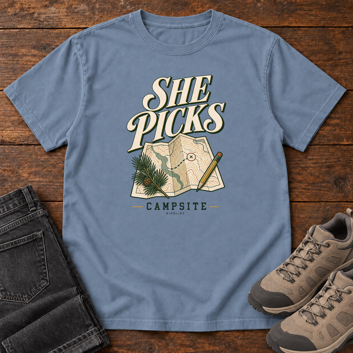 Blue Jean She Picks the Campsite T-Shirt with cream lettering, a folded map, pencil and evergreen sprig, front flat lay on wood.