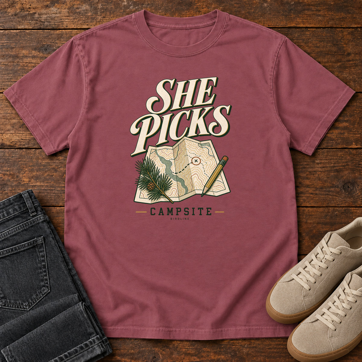 Brick She Picks the Campsite T-Shirt with cream lettering, a folded map, pencil and evergreen sprig, front flat lay on wood.