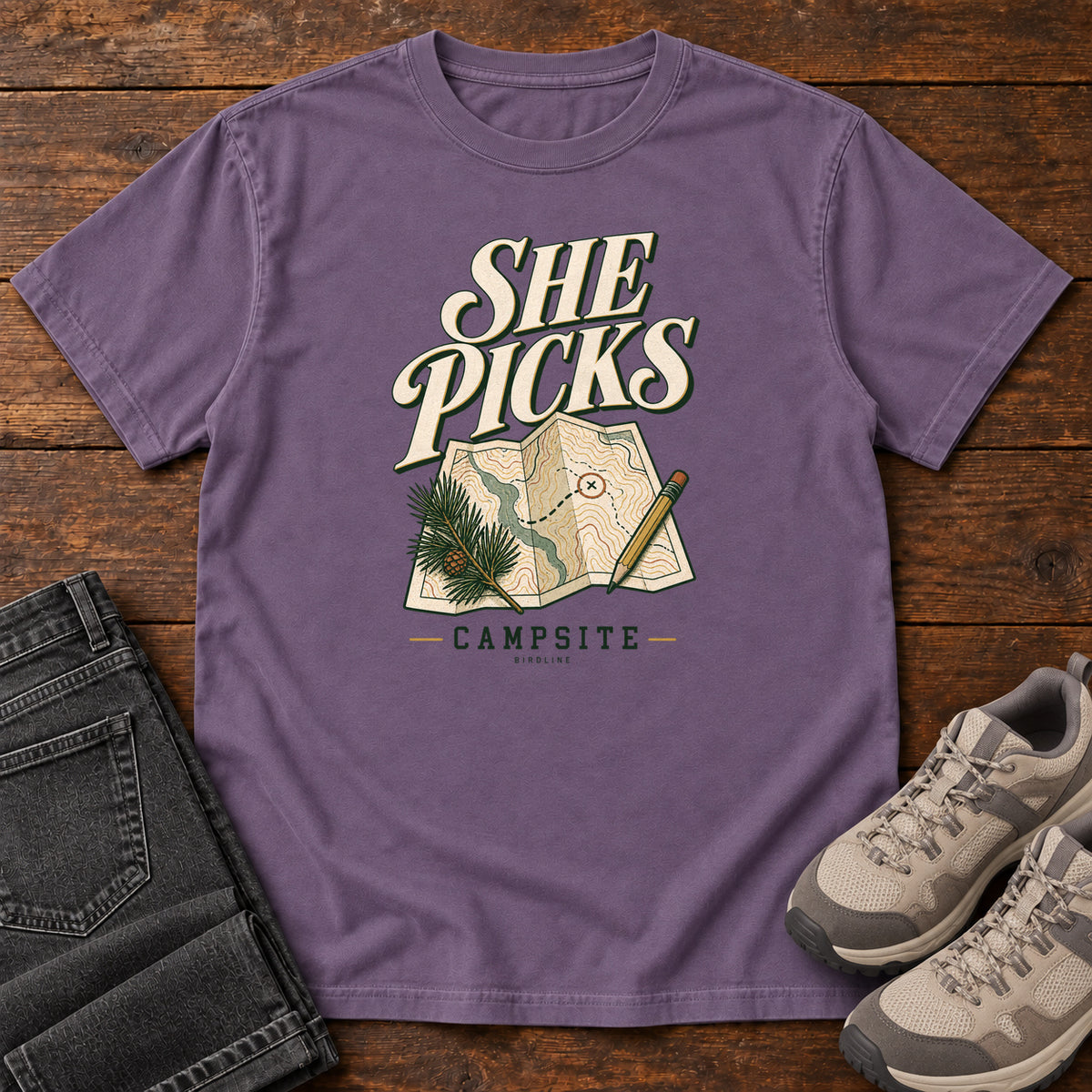Grape She Picks the Campsite T-Shirt with cream lettering, a folded map, pencil and evergreen sprig, front flat lay on wood.