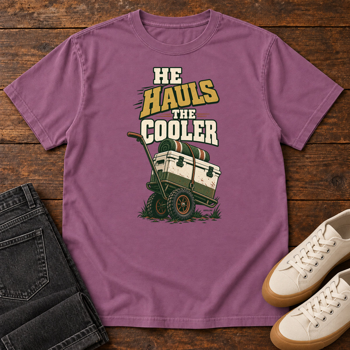 Berry He Hauls the Cooler T-Shirt with a cooler on a two-wheeled cart beneath He Hauls the Cooler lettering; front flat lay on wood.