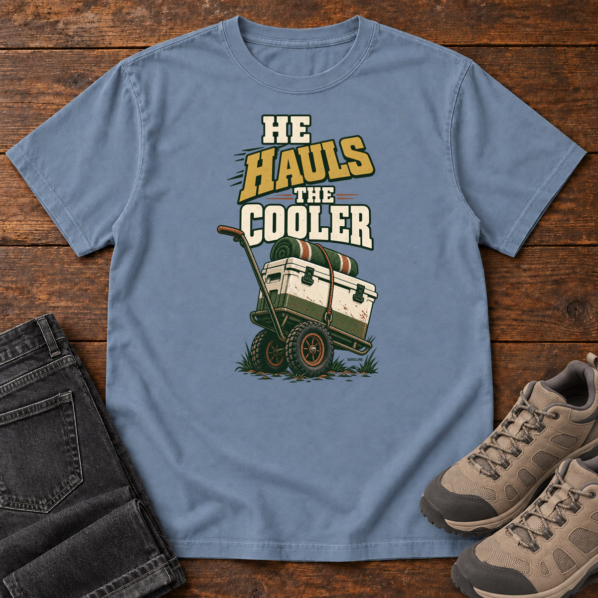 Blue Jean He Hauls the Cooler T-Shirt with a cooler on a two-wheeled cart beneath He Hauls the Cooler lettering; front flat lay on wood.