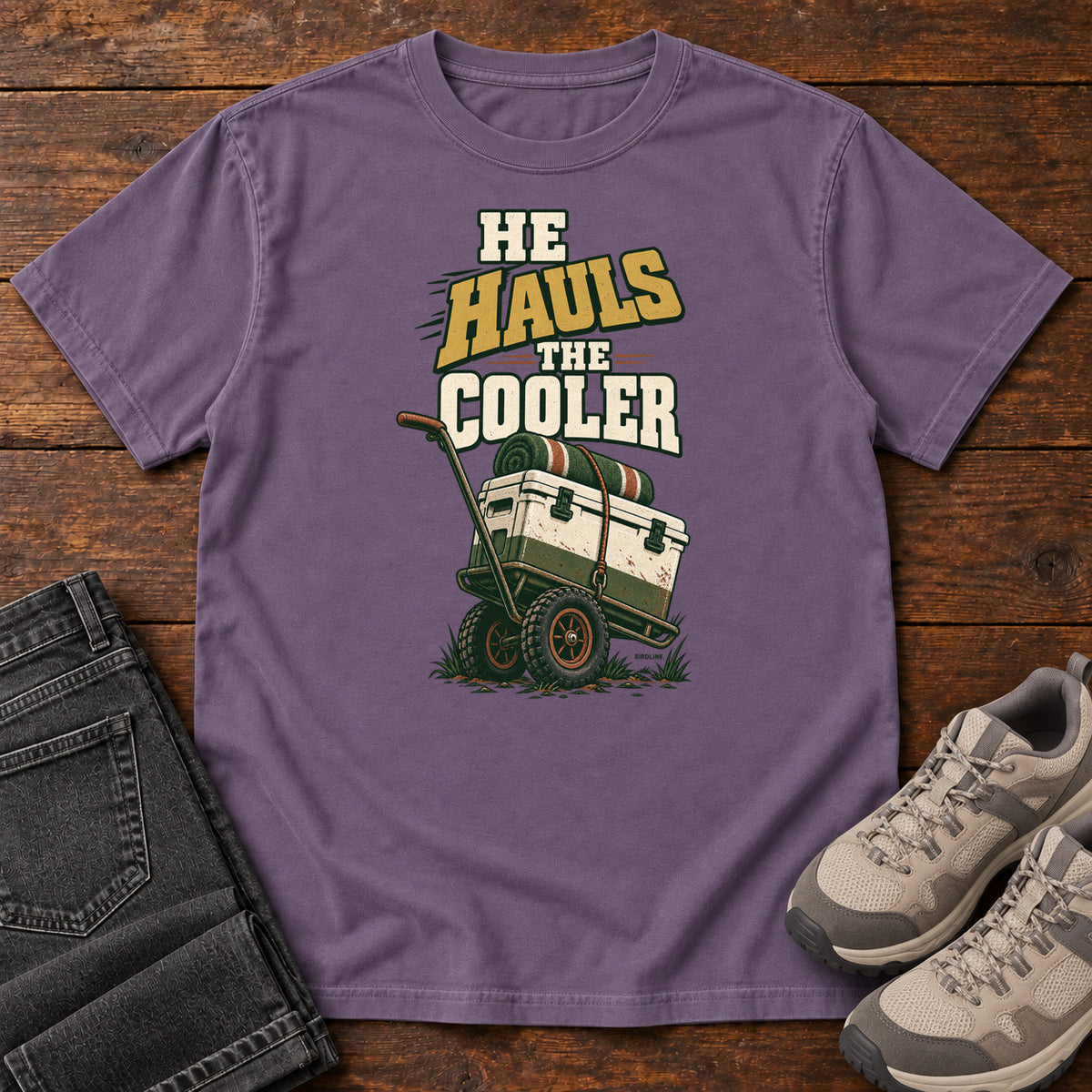 Grape He Hauls the Cooler T-Shirt with a cooler on a two-wheeled cart beneath He Hauls the Cooler lettering; front flat lay on wood.