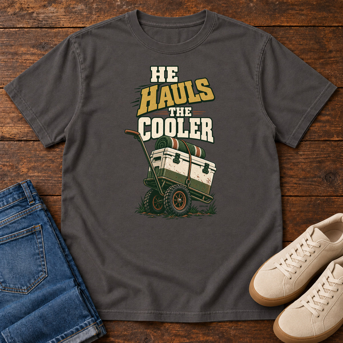 Graphite He Hauls the Cooler T-Shirt with a cooler on a two-wheeled cart beneath He Hauls the Cooler lettering; front flat lay on wood.