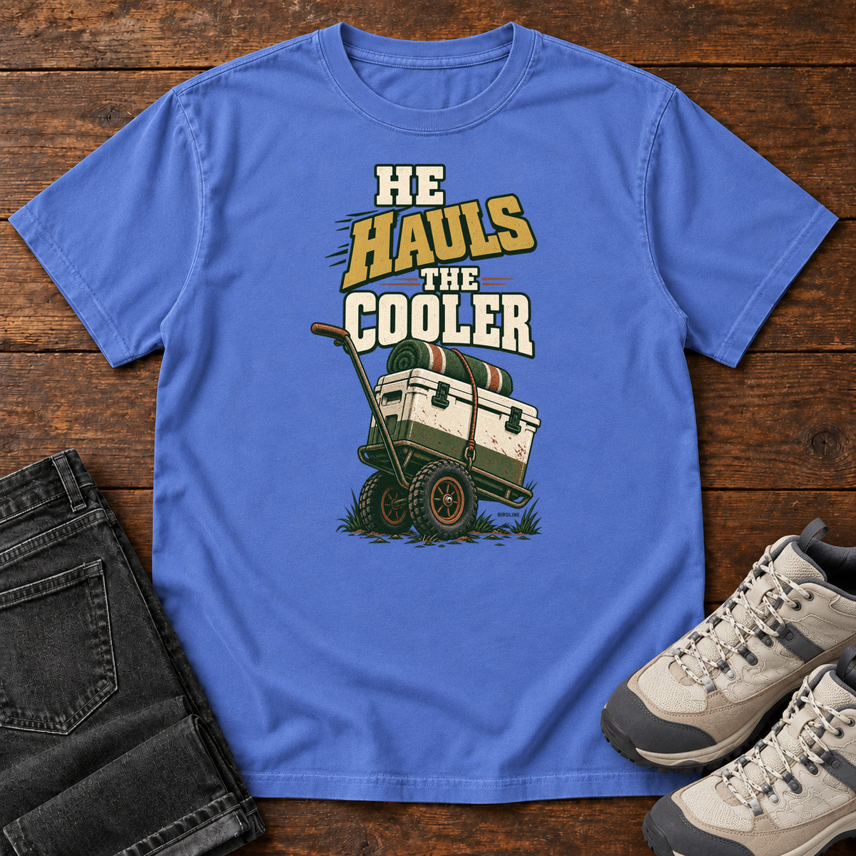 Mystic Blue He Hauls the Cooler T-Shirt with a cooler on a two-wheeled cart beneath He Hauls the Cooler lettering; front flat lay on wood.