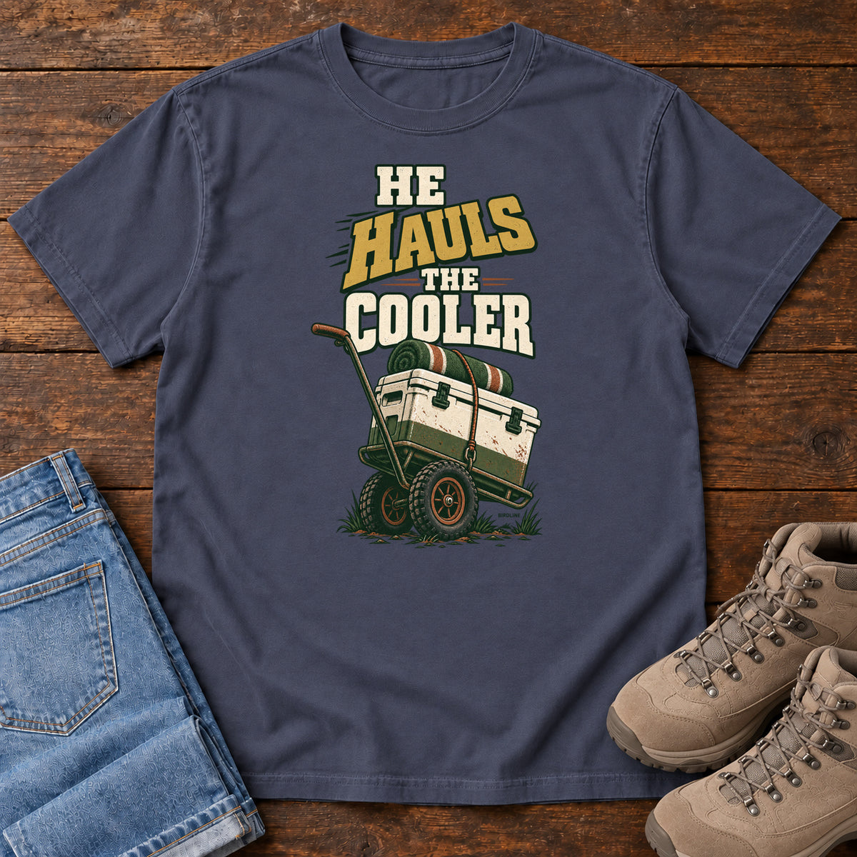 Navy He Hauls the Cooler T-Shirt with a cooler on a two-wheeled cart beneath He Hauls the Cooler lettering; front flat lay on wood.