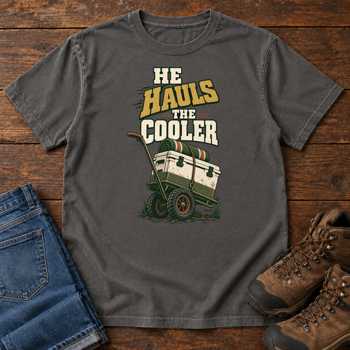 Pepper He Hauls the Cooler T-Shirt with a cooler on a two-wheeled cart beneath He Hauls the Cooler lettering; front flat lay on wood.