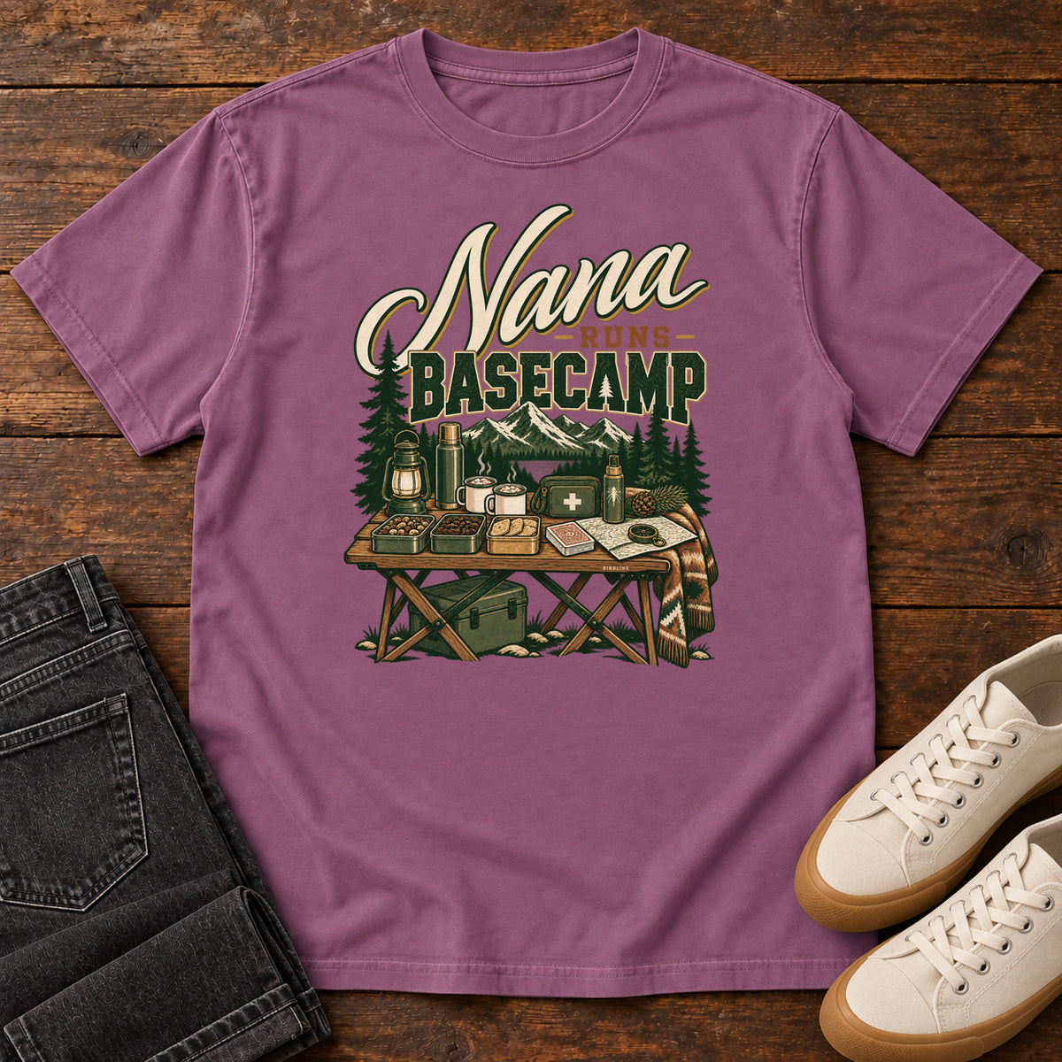 Berry Nana Runs Basecamp T-Shirt with a camp table with mugs, lantern, snacks and supplies beneath mountains; front flat lay on wood.