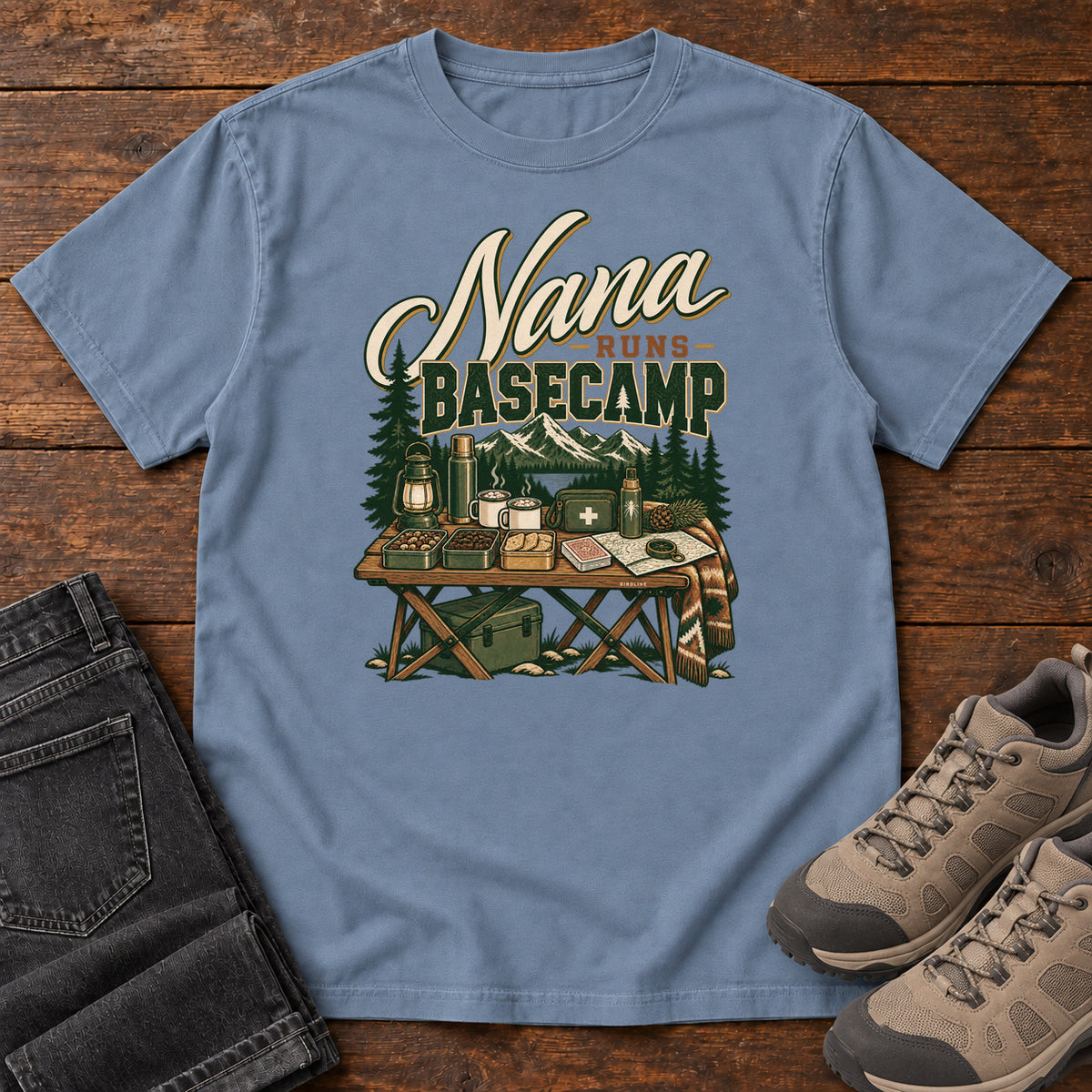 Blue Jean Nana Runs Basecamp T-Shirt with a camp table with mugs, lantern, snacks and supplies beneath mountains; front flat lay on wood.
